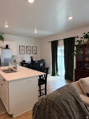 Open concept living area featuring a kitchen island with dual basin sink and white countertop, light wood-finish flooring, recessed lighting, and a sliding glass door with dark drapery