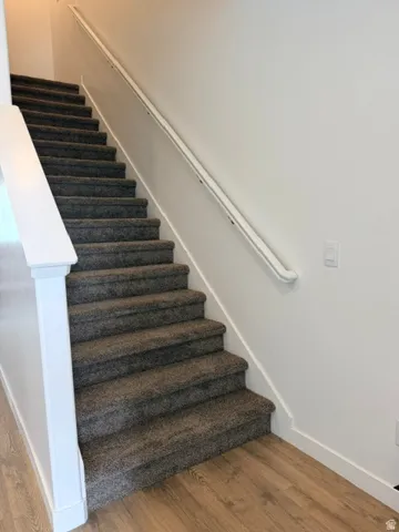 Carpeted staircase featuring a painted handrail and white newel post, complementing the wood-finish flooring and white baseboards