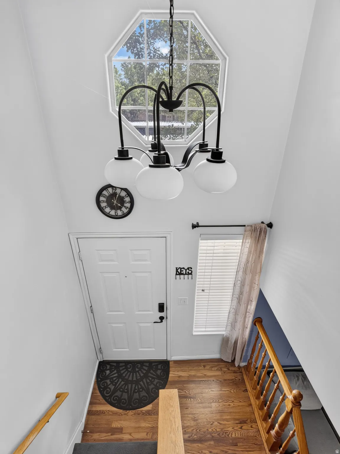Octagonal window with white trim, 5-light black chandelier, wood-finish flooring, white paneled door, and wood banister