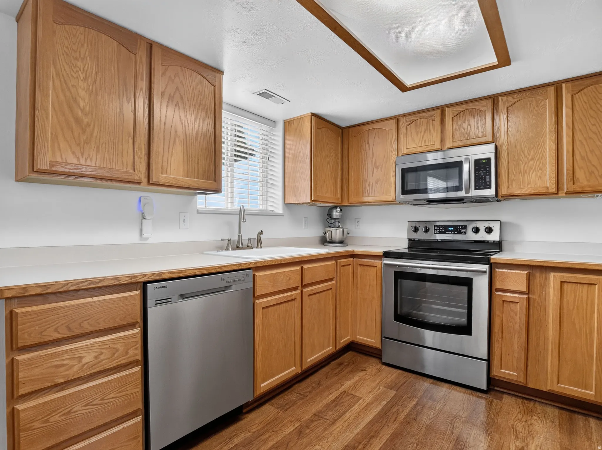 Kitchen featuring warm wood cabinetry, stainless steel appliances, a double basin sink, light-toned countertops, and wood-finish flooring