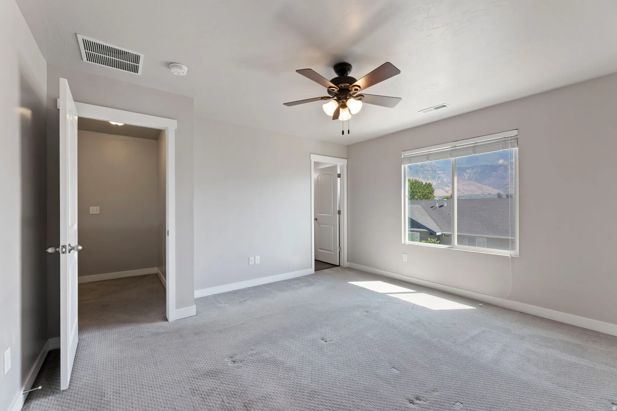 Spacious bedroom featuring light gray wall paint, textured gray carpeting, a multi-speed ceiling fan with integrated lighting, and a large window with white blinds