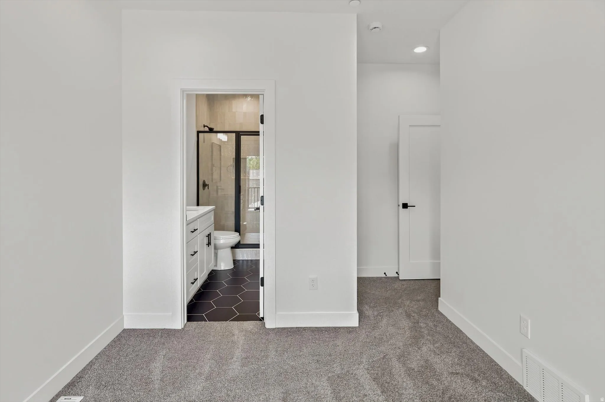 Bathroom entry featuring a vanity with shaker-style cabinetry, dark hexagonal floor tiles, a toilet, and a glass-enclosed shower with black framing
