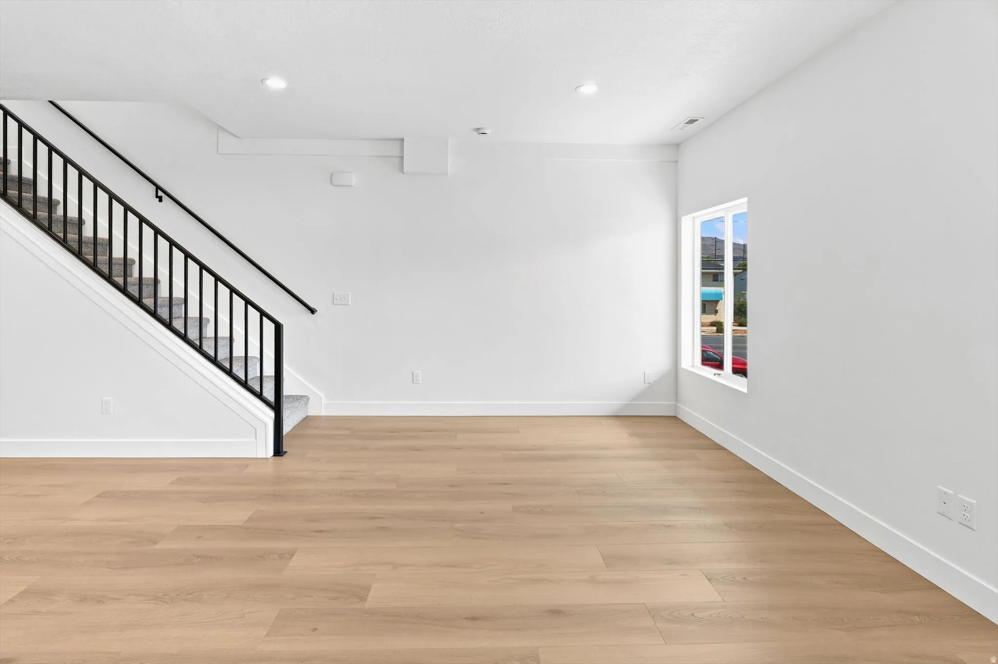 Spacious room featuring wood-finish flooring, a carpeted staircase with a contemporary black metal railing, and a window providing natural illumination