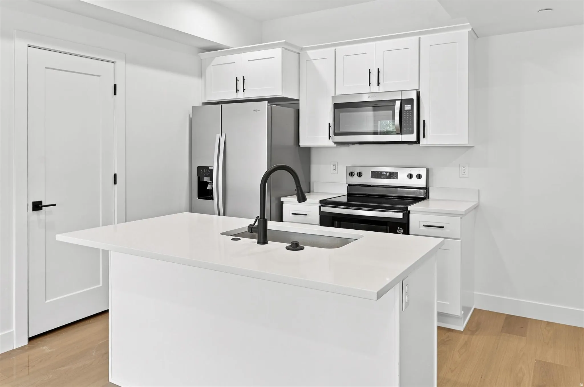 Modern kitchen featuring an island with an integrated sink and matte black faucet, stainless steel appliances, white cabinetry with black hardware, and wood-finish flooring