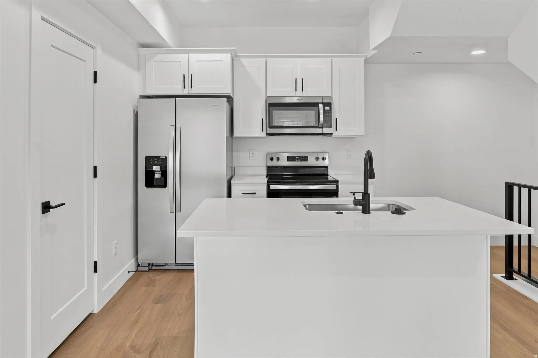Modern kitchen featuring white shaker cabinetry, stainless steel appliances, a center island with an integrated sink, black matte fixtures, and wood-finish flooring