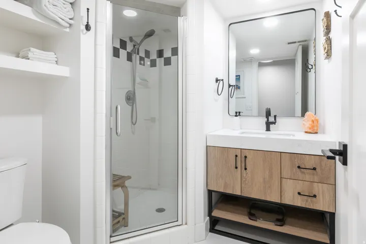 Modern bathroom featuring a glass-enclosed shower with black and white accent tiles, a vanity with wood-finish cabinetry, a white countertop, and a rectangular black-framed mirror