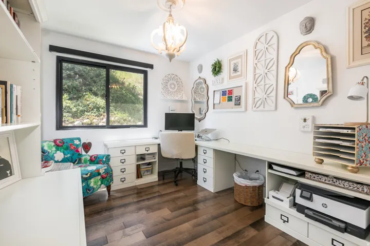 Dedicated workspace featuring wood-finish flooring, a large window with exterior views, built-in shelving, and extensive white desk cabinetry