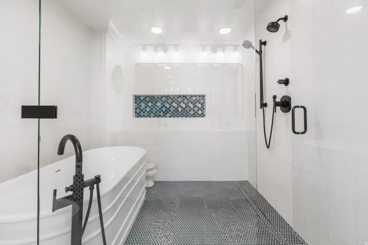 Spacious bathroom featuring a freestanding tub with a floor-mounted faucet, a walk-in shower with matte black fixtures, a tiled shower niche with decorative accent tiles, white subway tile walls, and a black and white penny tile floor