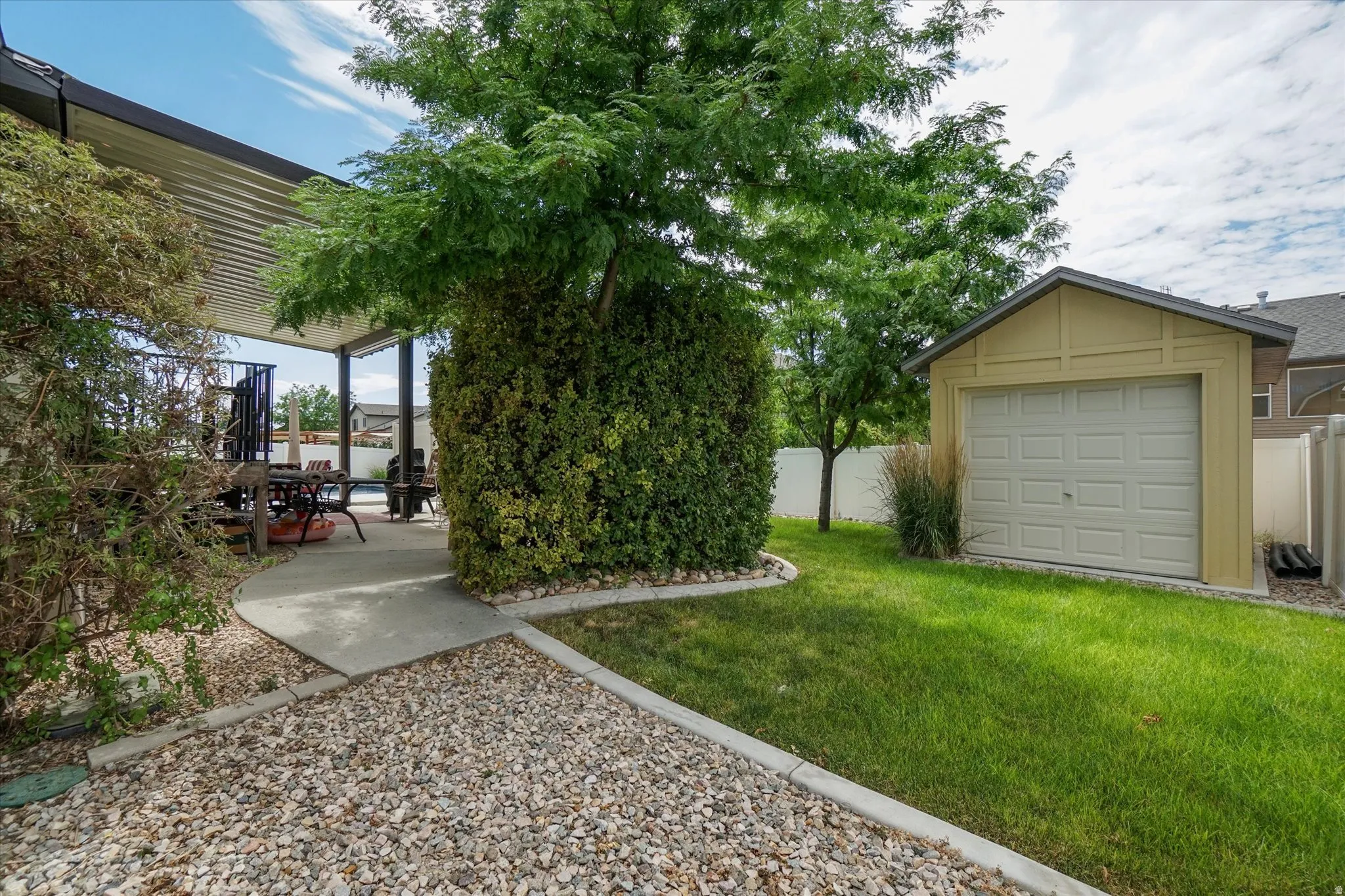 Expansive backyard featuring a covered patio with louvered roof, concrete pathway, gravel landscaping, manicured lawn, and a detached storage shed