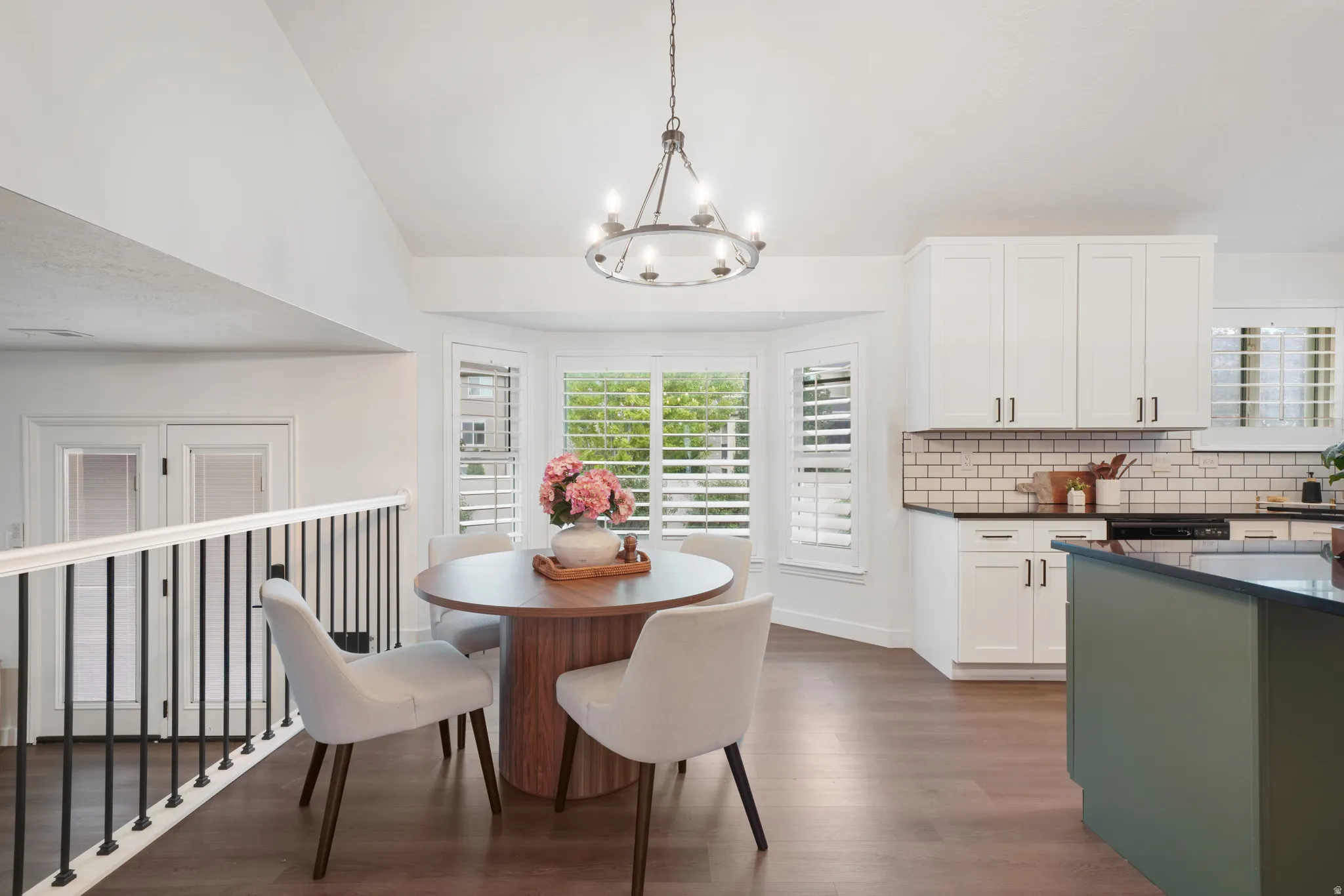 Open-concept kitchen and dining area featuring wood-finish flooring, a bay window with plantation shutters, white shaker cabinetry with dark hardware, a subway tile backsplash, and a central island with a dark countertop