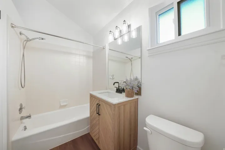 Bathroom featuring a contemporary wood-finish vanity with a single sink and black faucet, a white bathtub with tile surround, and wood-finish flooring