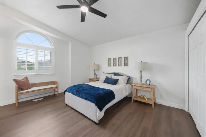 Bright bedroom featuring a large arched window with white plantation shutters, dark wood-finish flooring, a contemporary ceiling fan, and white walls