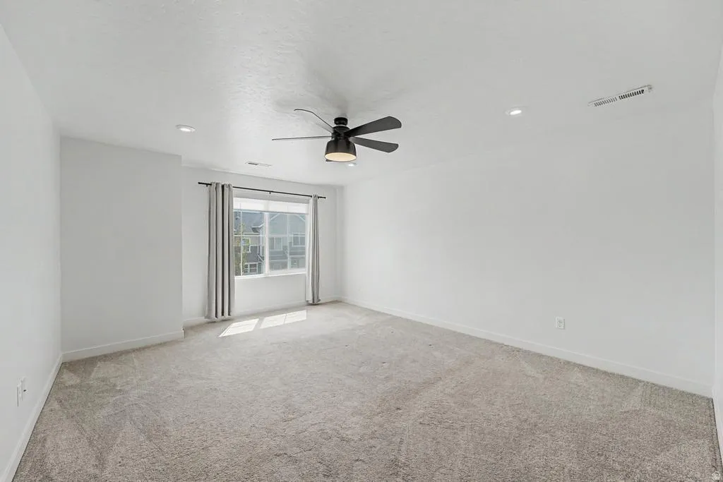 Spacious room featuring light gray carpeting, neutral white walls, a large window with gray drapes, an overhead ceiling fan with integrated lighting, and recessed lighting