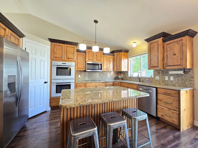 Kitchen featuring a central island with natural wood paneling, granite countertops, wood-finish flooring, stainless steel appliances including a double oven, and wood cabinetry with dark crown molding