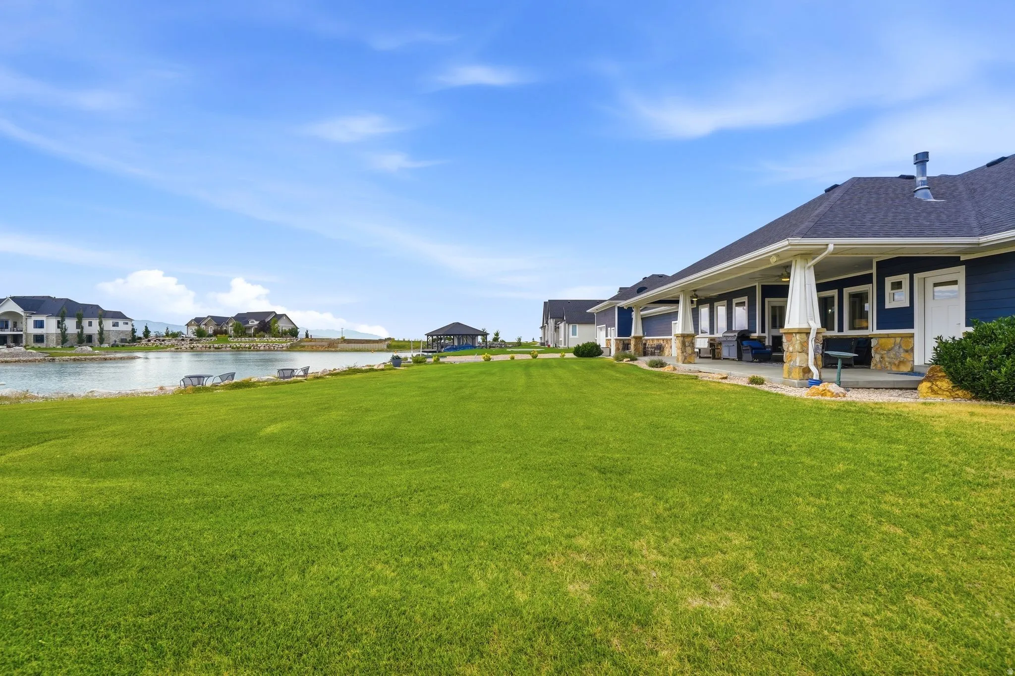 Expansive green lawn bordering a water feature, with a covered patio featuring stone-clad pillars and dark siding