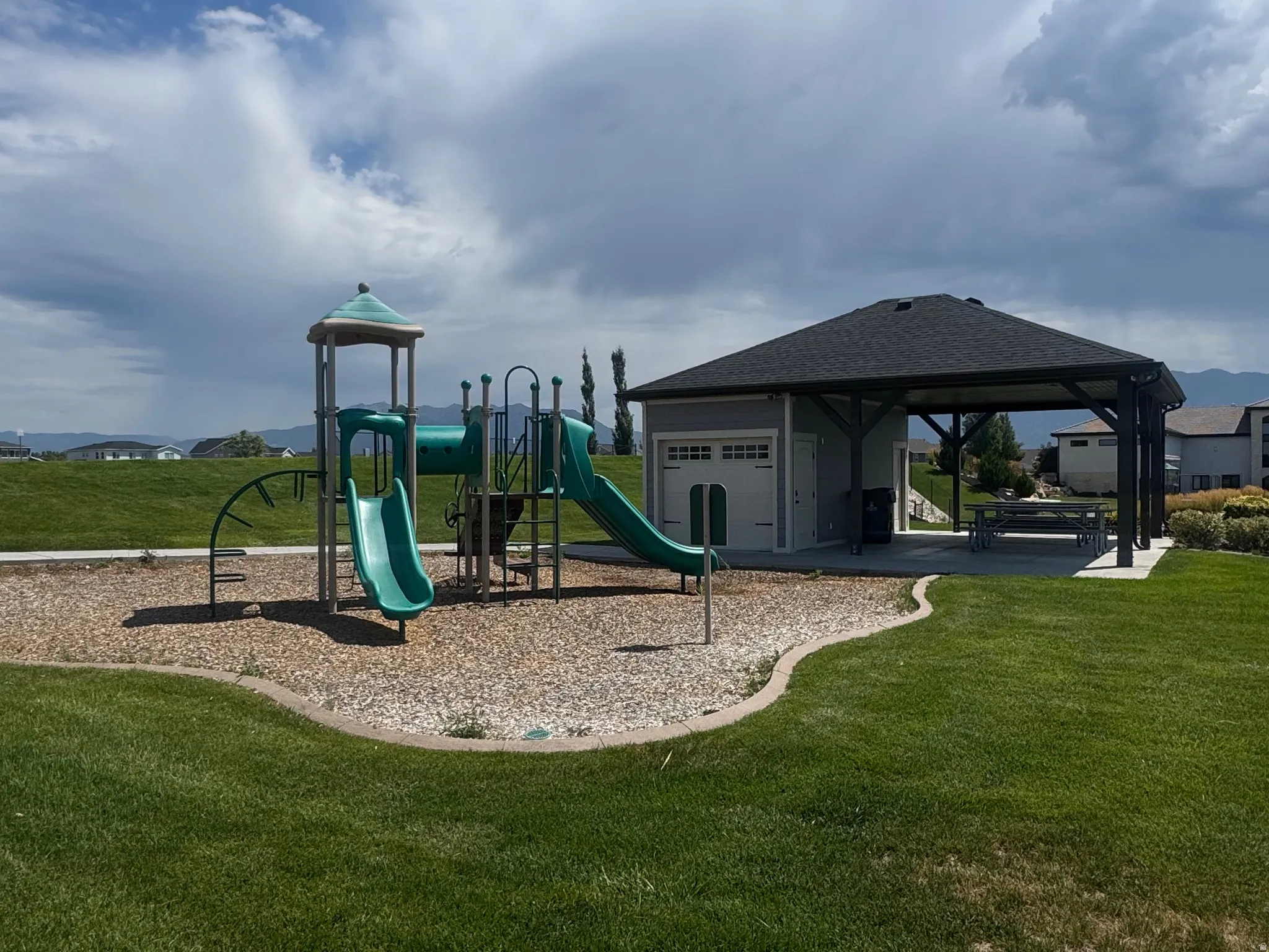 Community playground and covered picnic area with a dark shingle roof and concrete pad