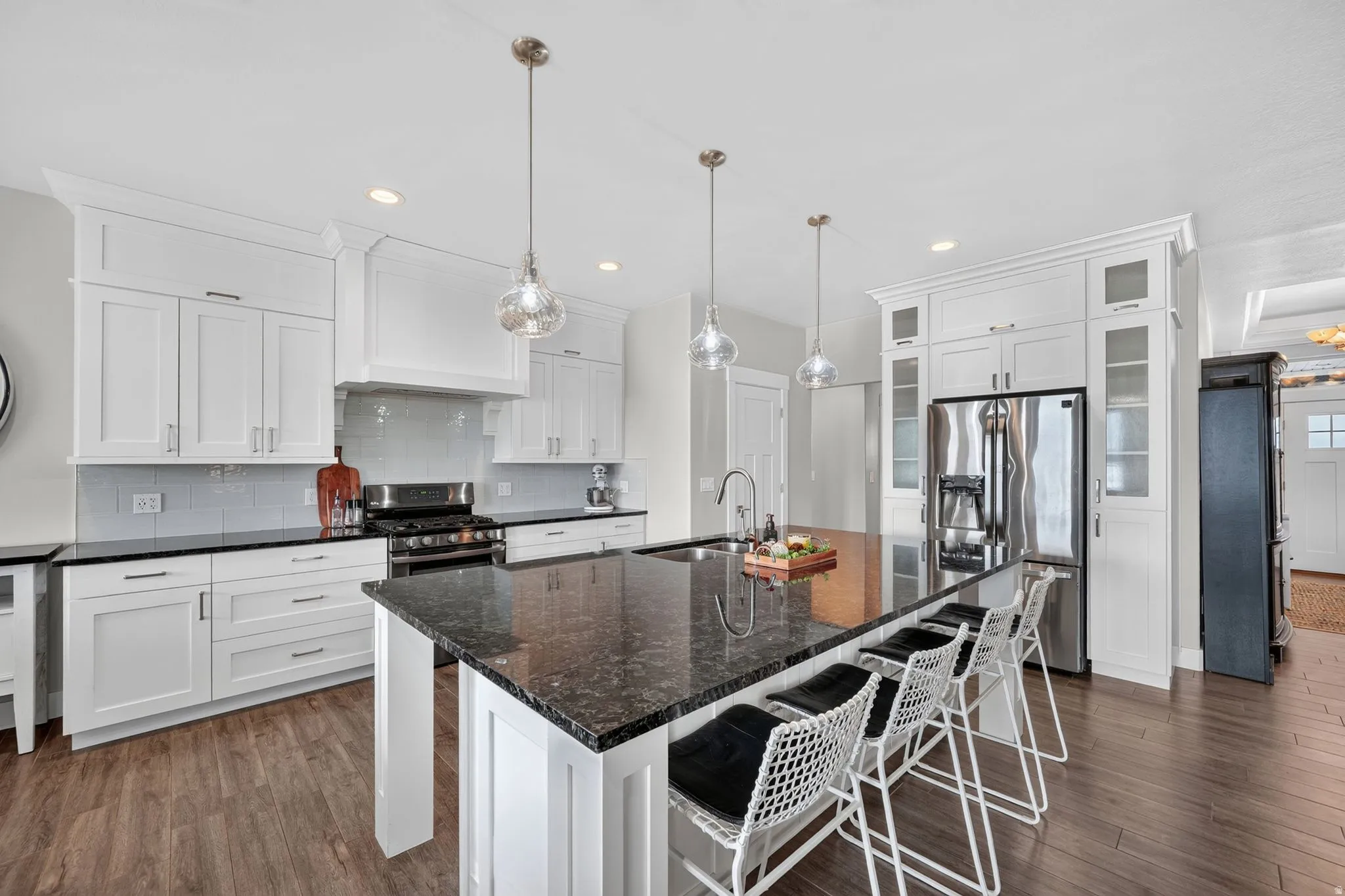 Spacious kitchen featuring an oversized island with seating, dark stone countertops, stainless steel appliances, white shaker-style cabinetry, and wood-finish flooring