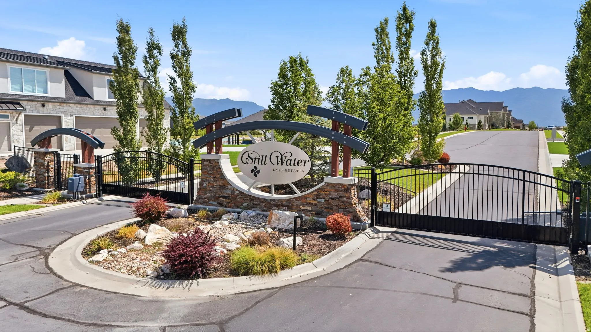 Gated community entry featuring a stone and metal sign, black wrought iron gates, and established landscaping