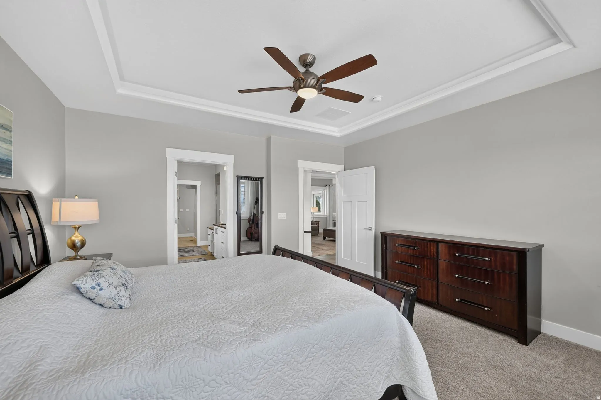 Recessed tray ceiling with a ceiling fan, light gray wall paint, white trim, and light-colored carpet flooring