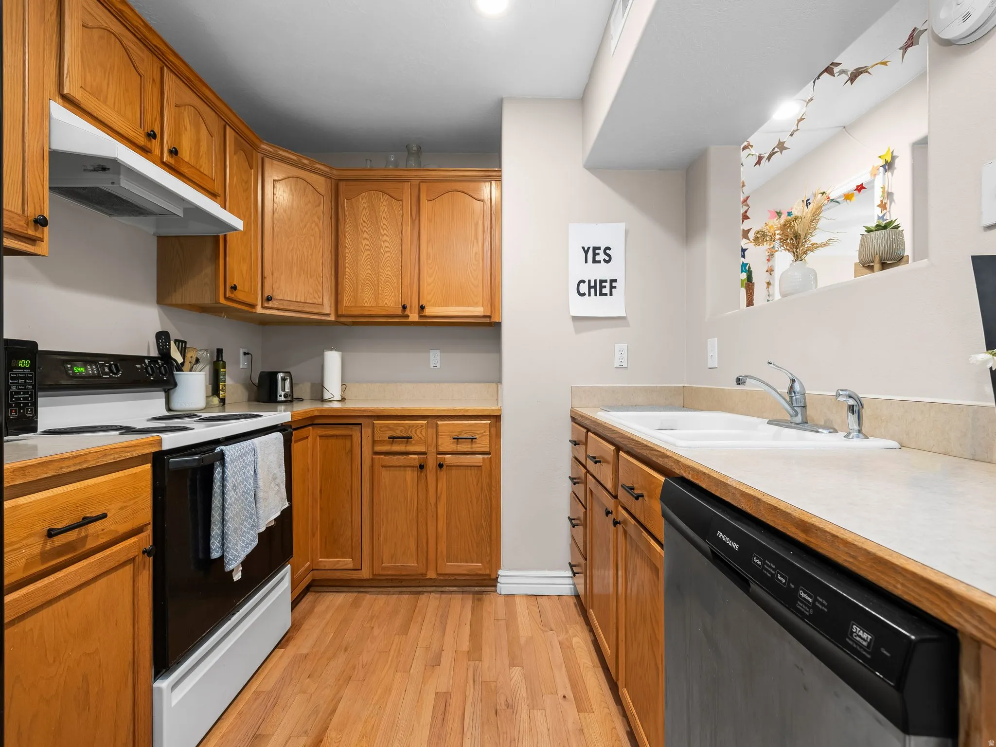 Kitchen featuring wood-finish flooring, natural wood cabinetry, light-toned countertops, a double basin sink, and a built-in dishwasher