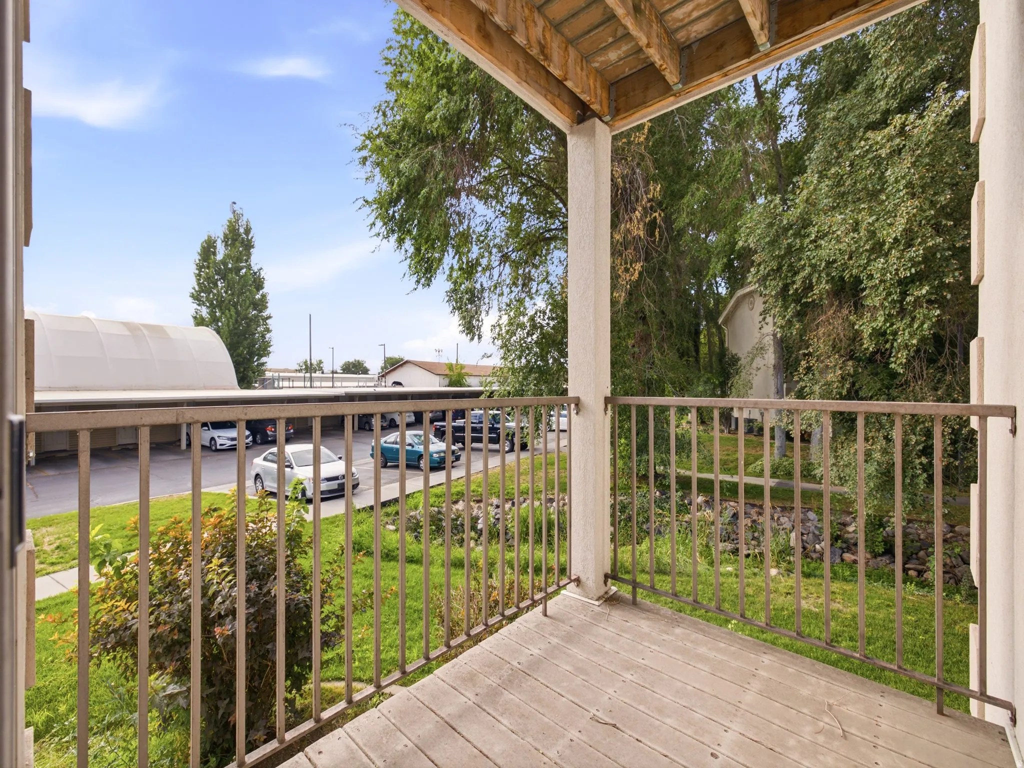 Balcony with wood-finish flooring and metal railings, overlooking a paved parking area and mature trees
