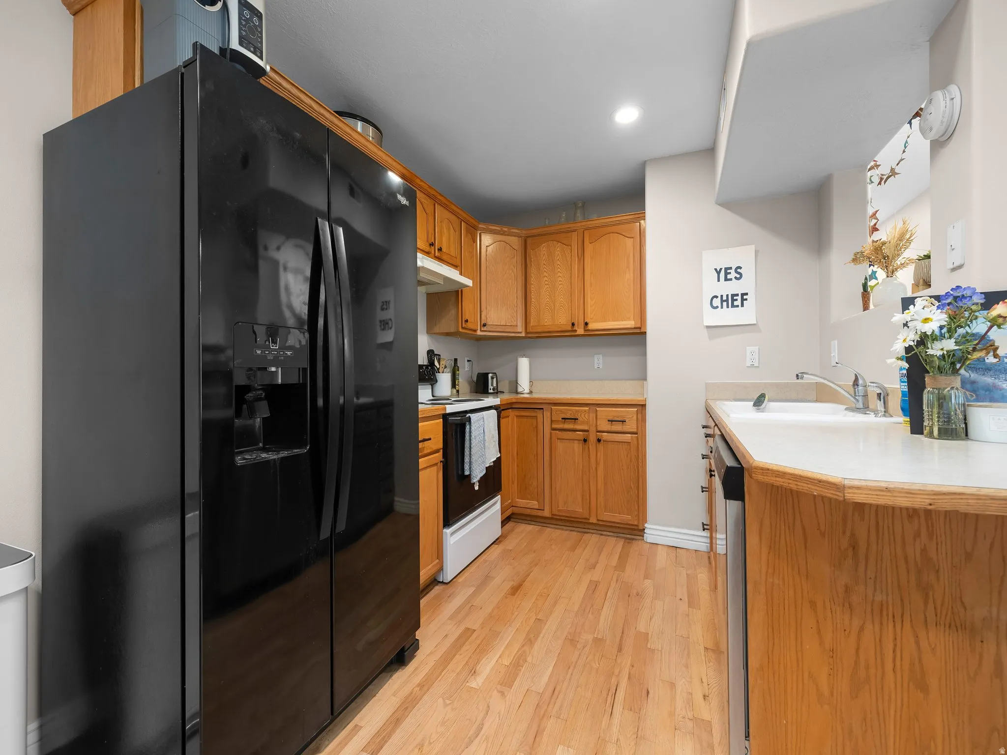 Kitchen featuring wood-finish flooring, natural wood cabinetry, a built-in stainless steel dishwasher, and a black side-by-side refrigerator