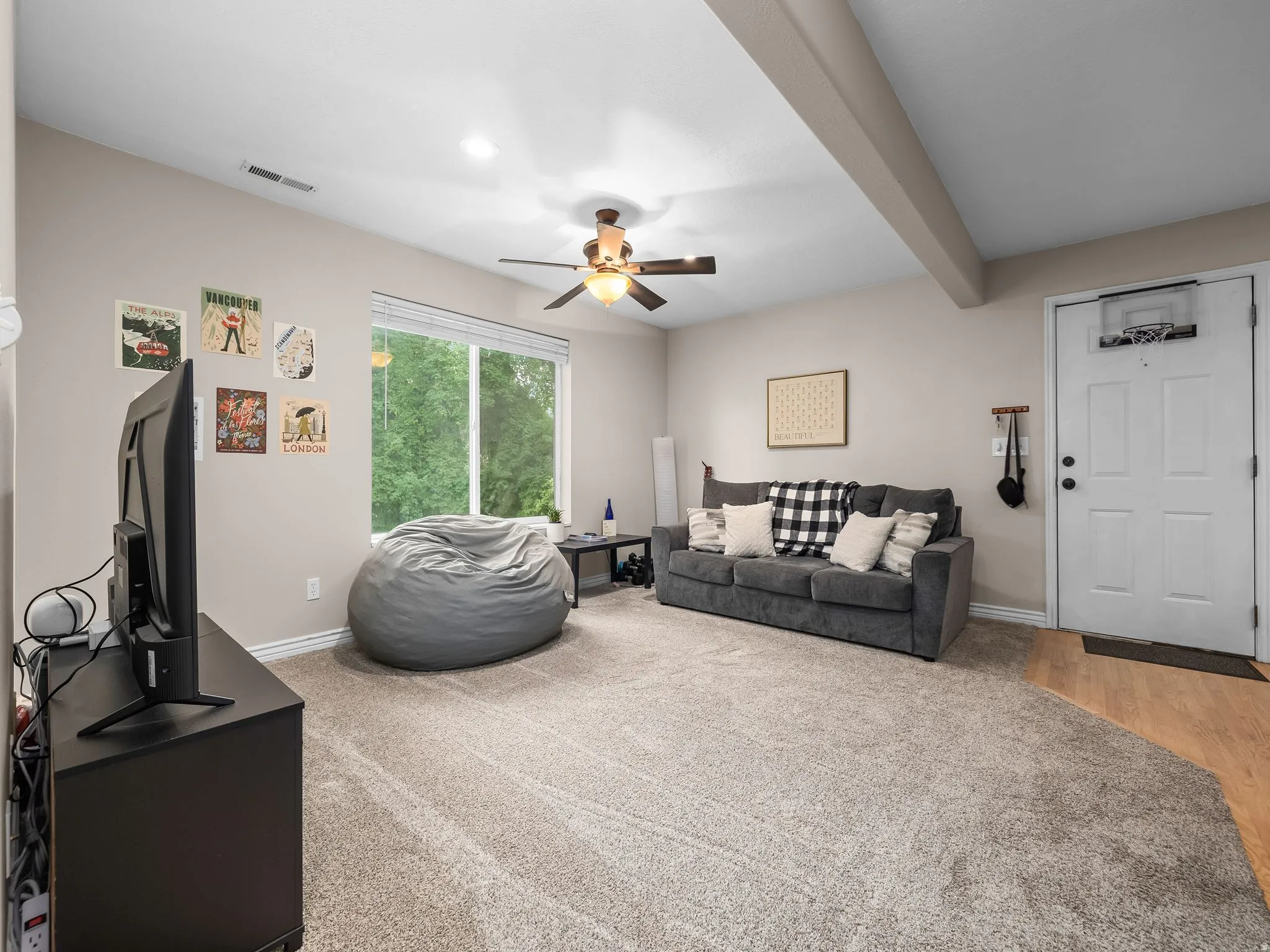 Carpeted living area featuring a large window, a ceiling fan, light-colored walls, and a visible wood-finish flooring section near the entry
