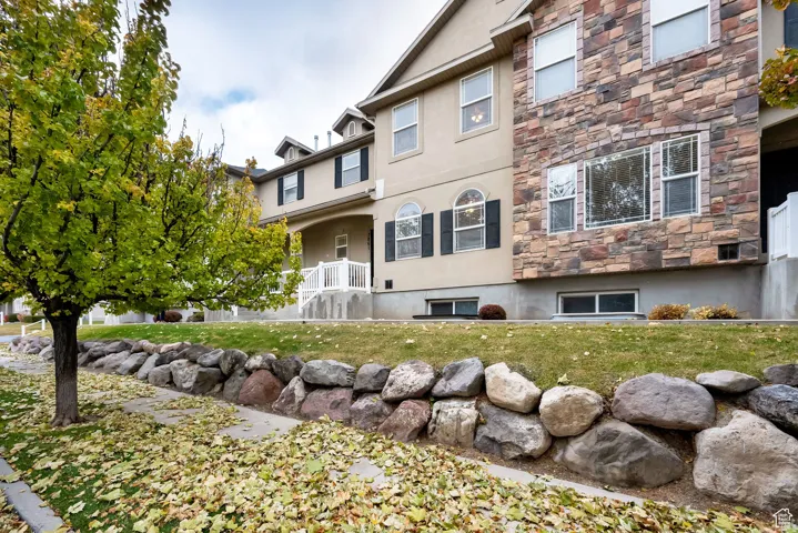 Contemporary multi-story exterior featuring a stone and stucco facade, a covered entry porch with white railings, and mature landscaping with a stone retaining wall