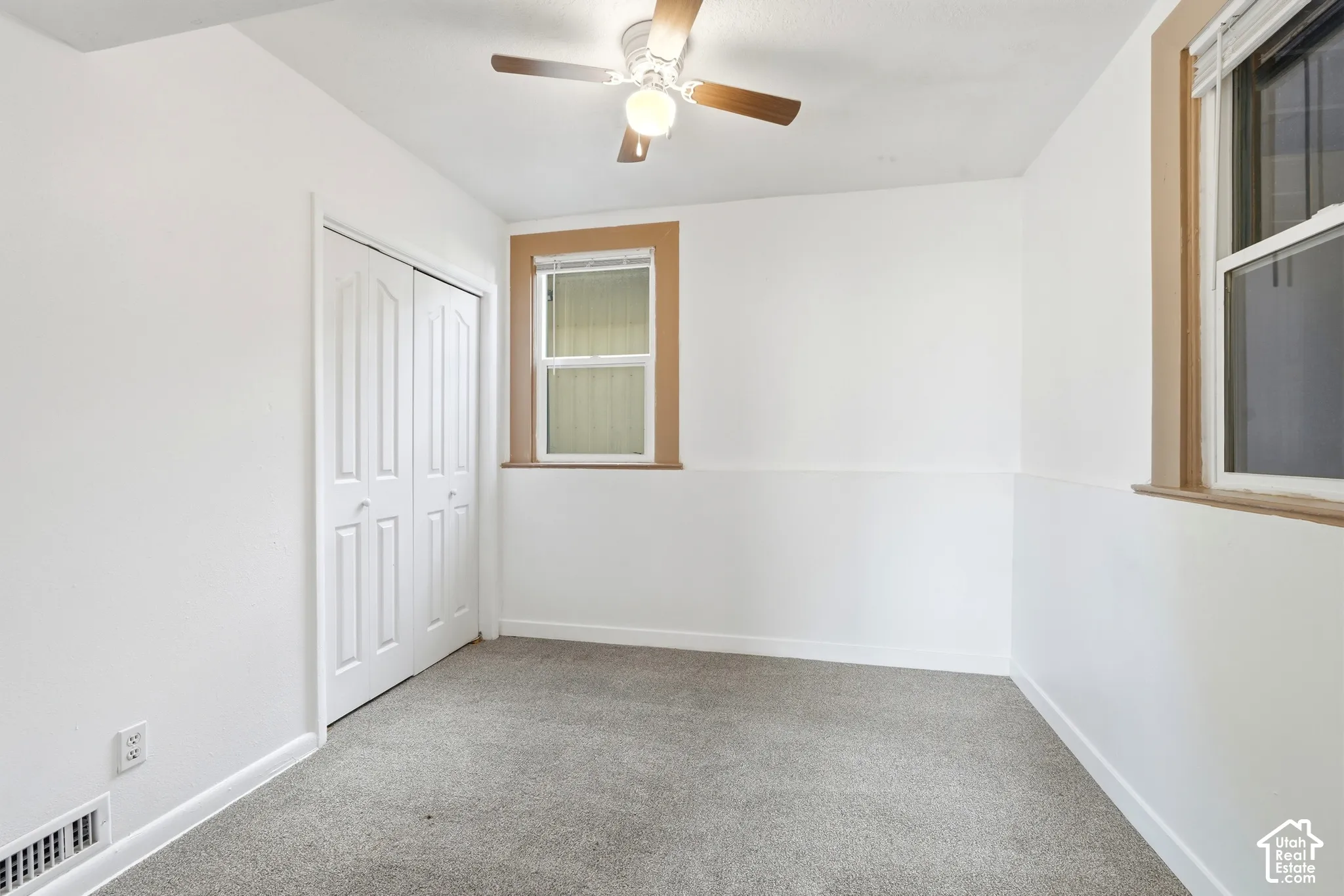 Carpeted room featuring a ceiling fan, two windows with wood-finish trim, white paneled walls, and a bi-fold closet door