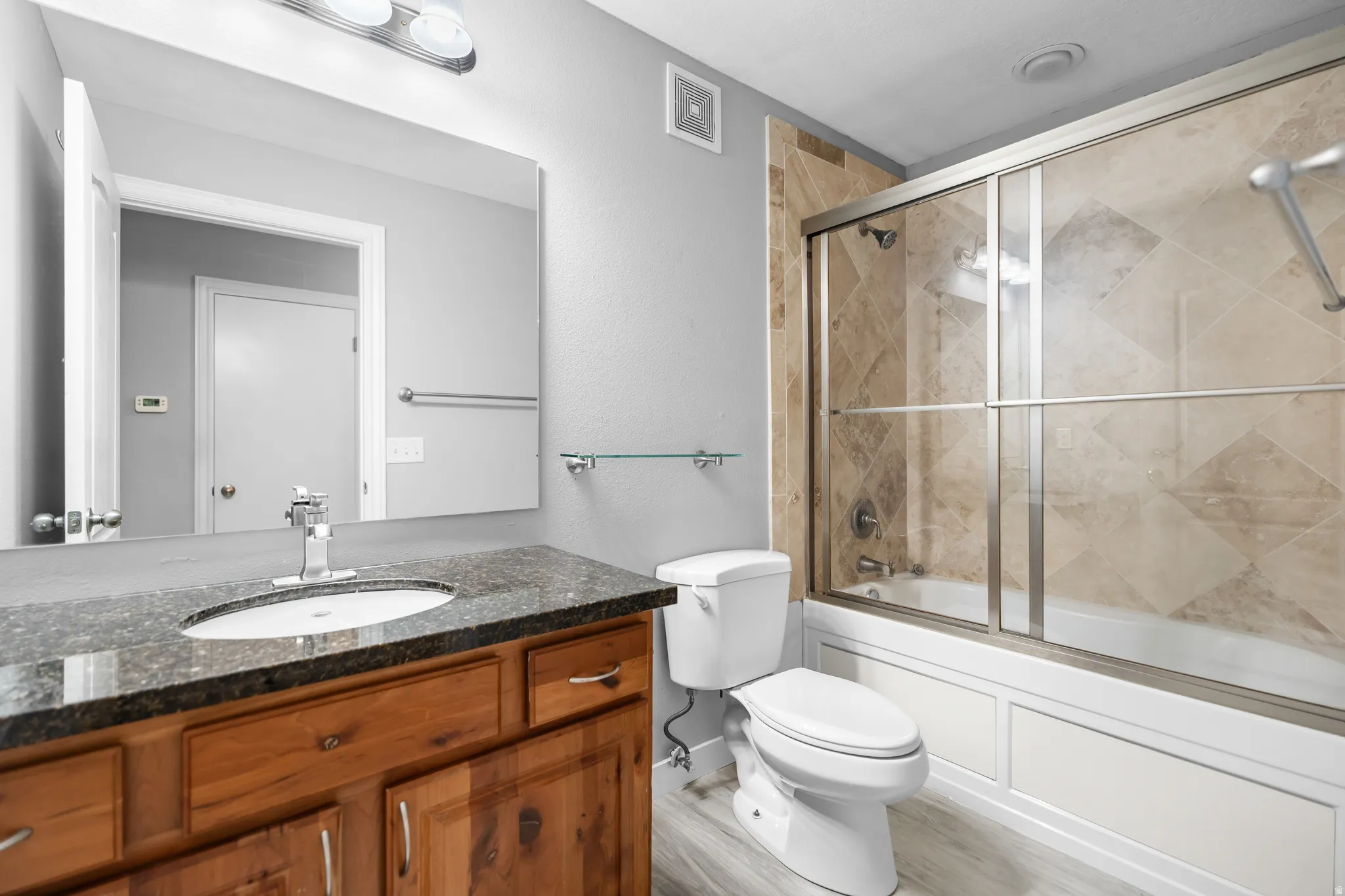 Bathroom featuring a wood-finish vanity with a dark speckled countertop and undermount sink, a large mirror, a tile-surround shower-tub combination with sliding glass doors, and light wood-finish flooring