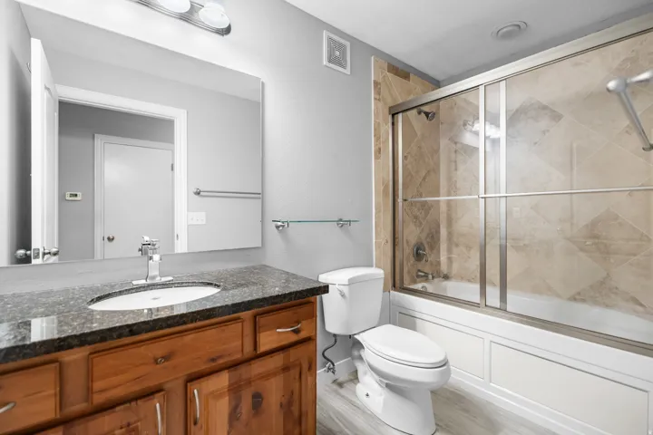 Bathroom featuring a wood-finish vanity with a dark speckled countertop and undermount sink, a large mirror, a tile-surround shower-tub combination with sliding glass doors, and light wood-finish flooring