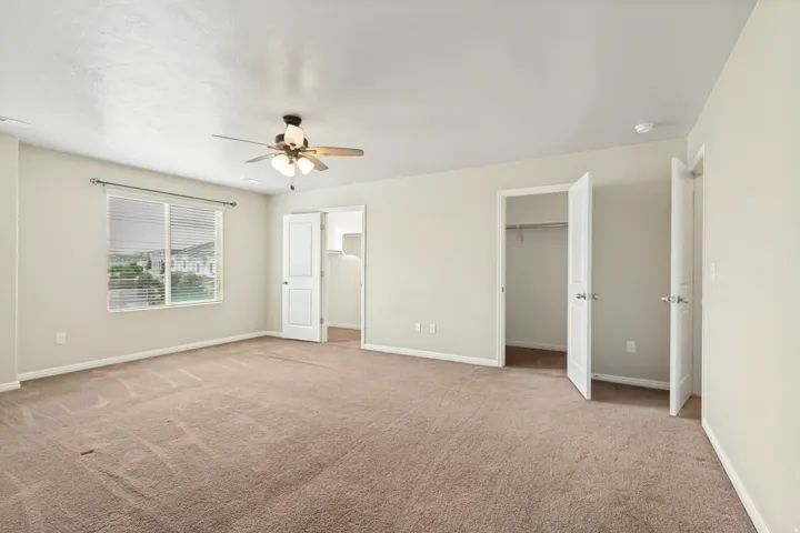 Spacious room featuring neutral carpet flooring, a ceiling fan with light fixture, and a window with blinds overlooking distant mountains and buildings