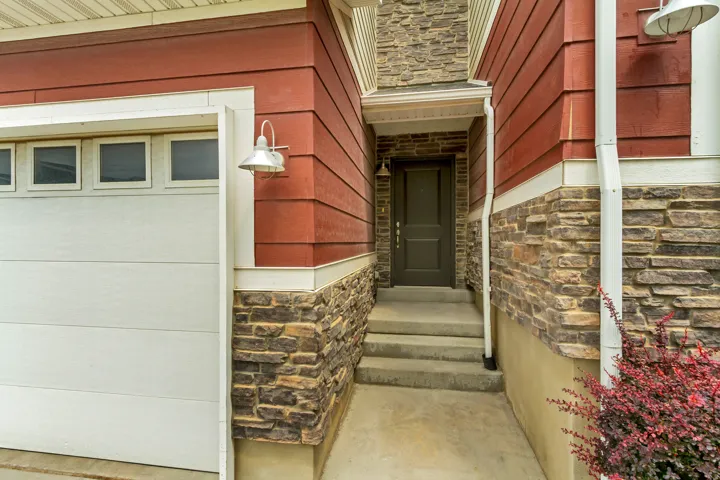 Exterior featuring red horizontal siding, stacked stone accents, a dark entry door with transom window, and a white garage door with rectangular window panels