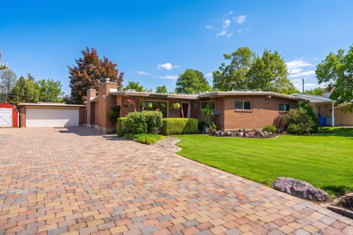 Brick exterior residence featuring a multi-color paver driveway, an attached two-car garage, a detached red storage shed, a manicured lawn, and mature landscaping