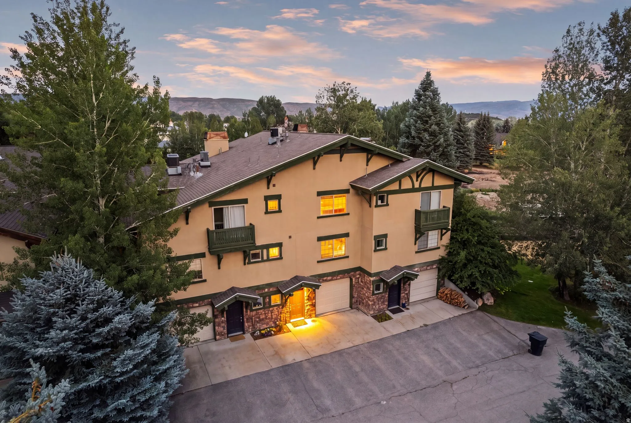 Multi-story stucco structure featuring a brown roof, stone accents, multiple balconies, and integrated garages, surrounded by mature trees