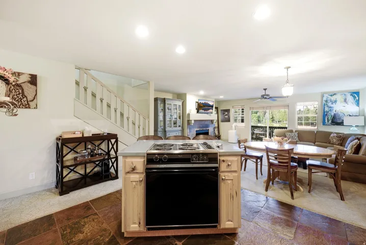 Open-concept living space featuring a central kitchen island with an integrated oven, stone tile flooring, a carpeted living area, a staircase with white balusters, and a sliding glass door providing access to an outdoor area