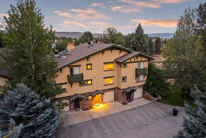 Multi-story stucco structure featuring a brown roof, stone accents, multiple balconies, and integrated garages, surrounded by mature trees