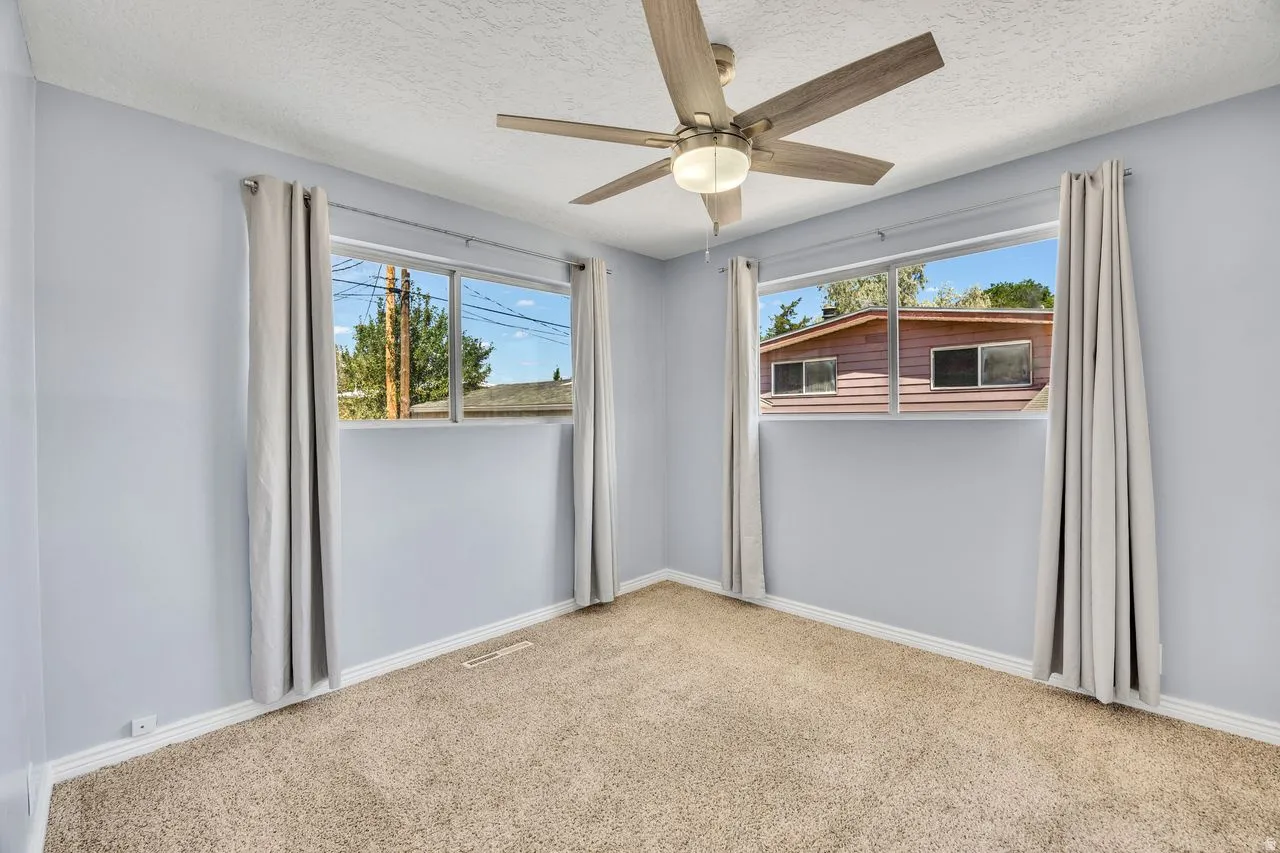 Carpeted room featuring light blue walls, two large windows with neutral drapes, and a ceiling fan with integrated lighting