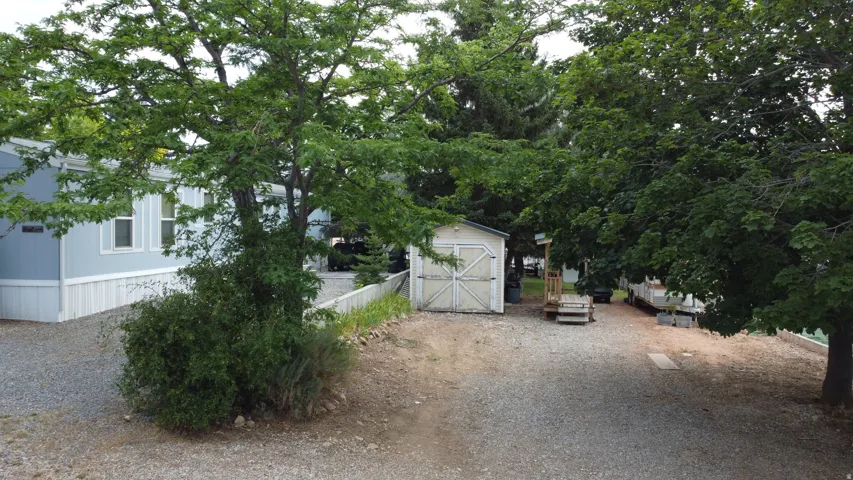 Spacious gravel yard featuring mature trees, a light blue-sided home with white trim, a storage shed, and a raised wooden deck