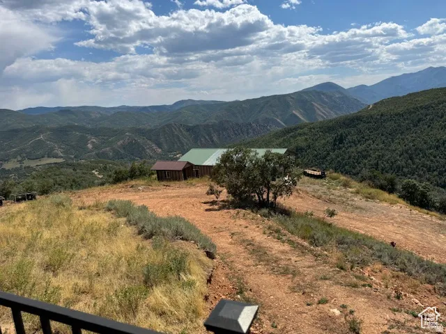 Expansive mountainous terrain with distant water feature, featuring a building with a green metal roof and rustic wood siding, surrounded by natural landscaping