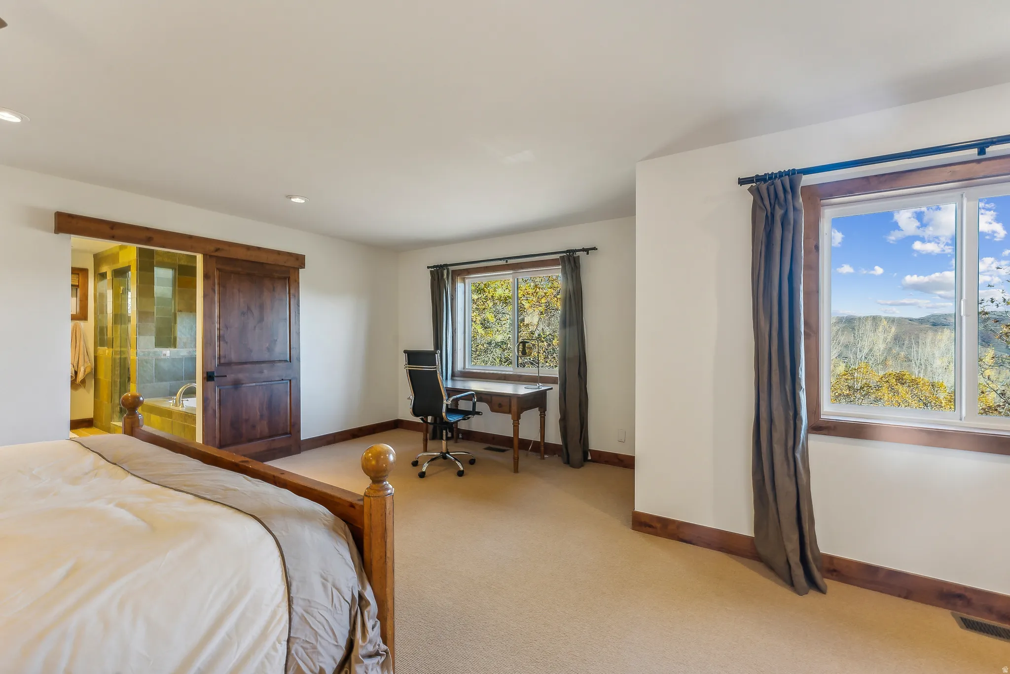 Carpeted bedroom featuring a large window with a distant landscape, wood-trimmed windows, and a sliding barn door leading to a bathroom with a tiled shower and bathtub