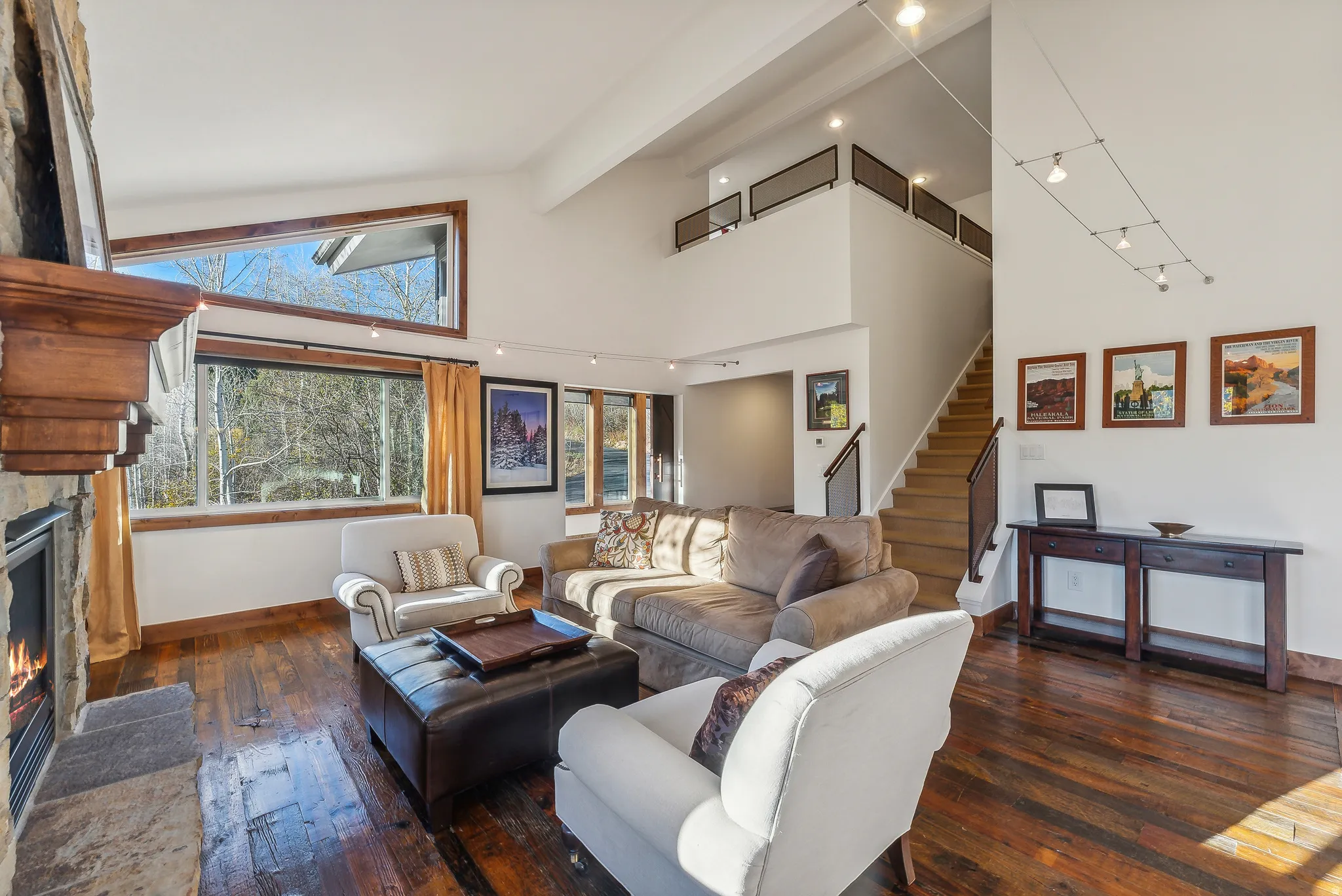 Spacious living area featuring a stone fireplace with a wood mantel, rich wood-finish flooring, high ceilings, large windows with wood trim, and a second-story interior balcony with metal railing