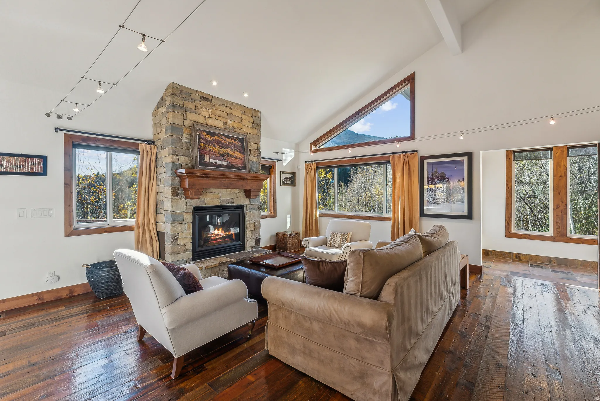 Vaulted ceiling living room featuring a stone-clad fireplace with a wood mantle, wood-finish flooring, and multiple large windows