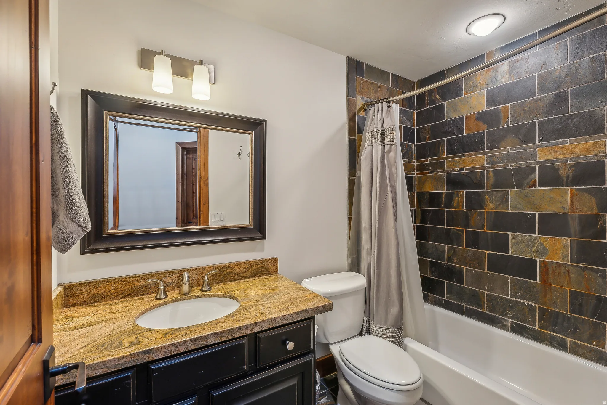 Bathroom featuring a tub/shower combination with a natural stone tile surround, a single vanity with a granite countertop, and a framed mirror with overhead lighting