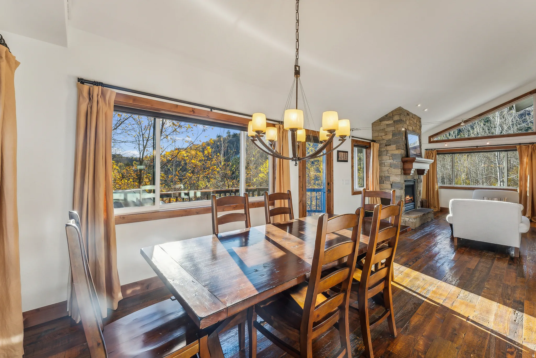 Open-concept living area featuring rich wood-finish flooring, a stone-clad fireplace, and expansive windows