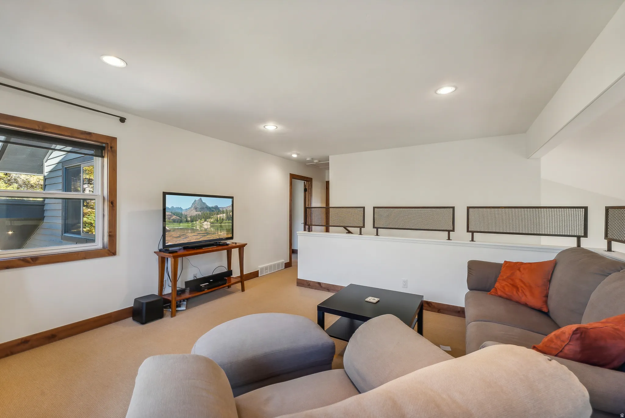 Spacious carpeted living area featuring recessed lighting, a window with wood trim, and a partial wall with decorative metal railing