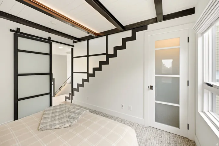 Bedroom featuring exposed ceiling beams, a sliding barn door with frosted glass panels, an open-tread staircase with metal framing, a multi-pane glass door, and a textured carpeted floor