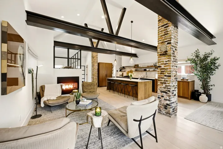 Open-concept living space featuring vaulted ceilings with exposed steel beams, a modern fireplace, light wood-finish flooring, a brick accent column, and a kitchen island with seating