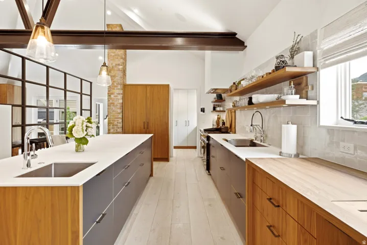 Expansive kitchen featuring light wood-finish flooring, a generous central island with an undermount sink and contemporary cabinetry, vaulted ceilings with exposed structural beams, a light-toned tile backsplash, and open shelving