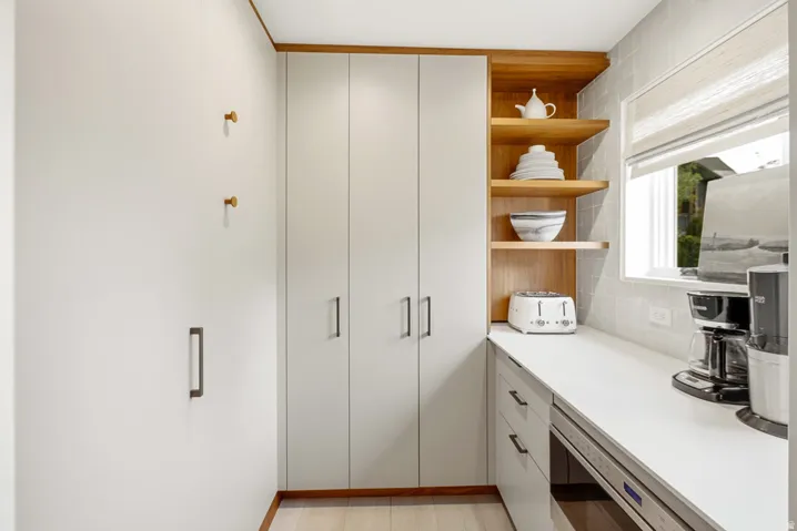 Modern pantry area featuring extensive built-in cabinetry with sleek hardware, light wood-finish open shelving, a broad countertop space, and a tiled backsplash